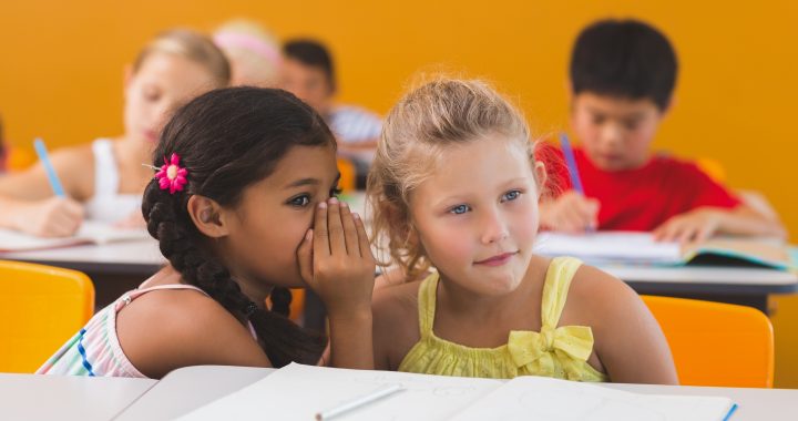 Schoolgirl whispering into her friend s ear in classroom